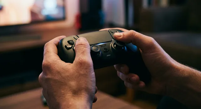 Manos sosteniendo un mando de videojuegos en un entorno en penumbra con reflejos de luz ambiental.